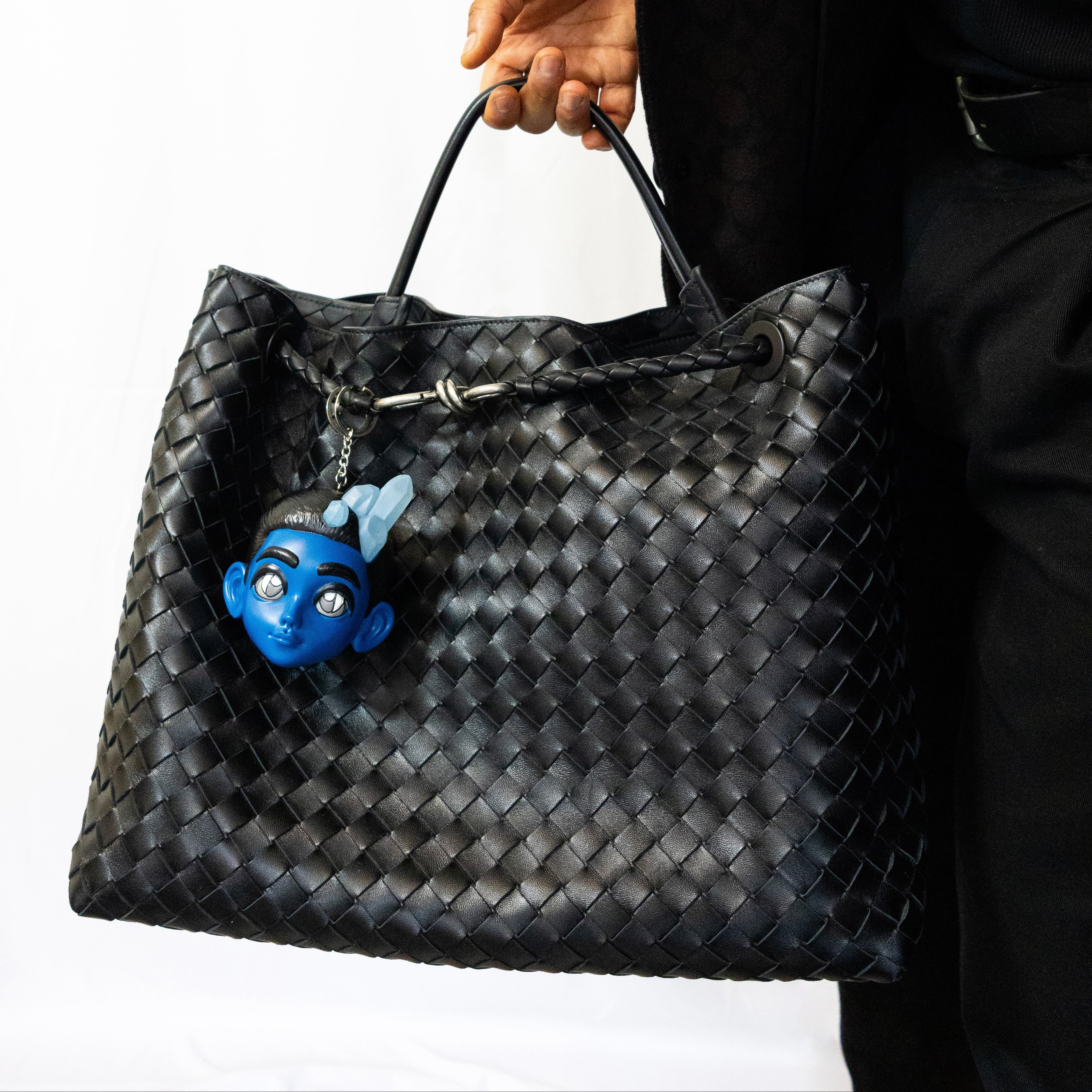 Person holding a black woven handbag with a blue character charm on a white background