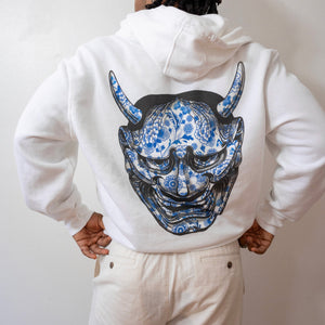 Person wearing a white hoodie with a blue and black mask design on the back against a plain background