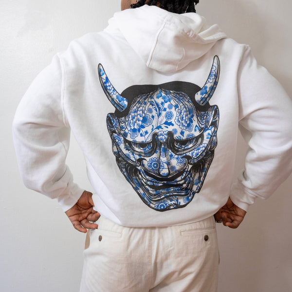 Person wearing a white hoodie with a blue and black mask design on the back against a plain background